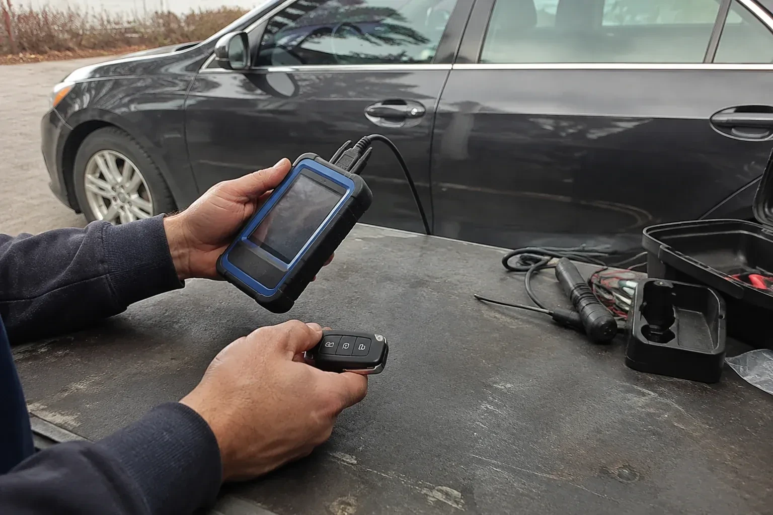 Hands holding a blue-edged electronic device and car-key fob at a worktable beside a parked car. — Transponder Key Programming, Mississauga